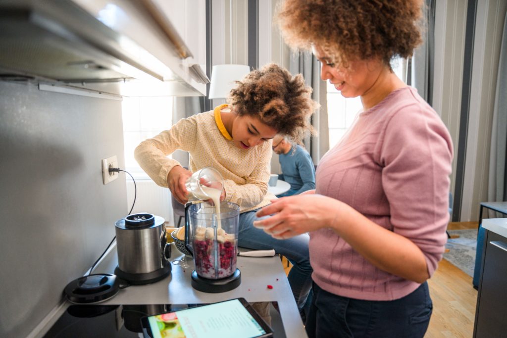Mother and daughter are preparing smoothie for smoothie bowl in a blender. They are putting raspberries and milk in a blender. They are using digital tablet and reading recipe.